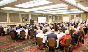 A room full of people sitting at tables watch a presenter speak from a stage.