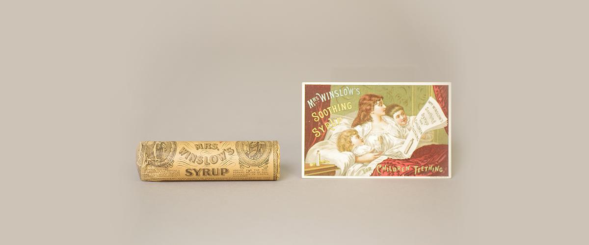   A medicine bottle wrapped in tan, printed paper next to a colorful card featuring a woman and her two children looking at a newspaper advertisement. 
