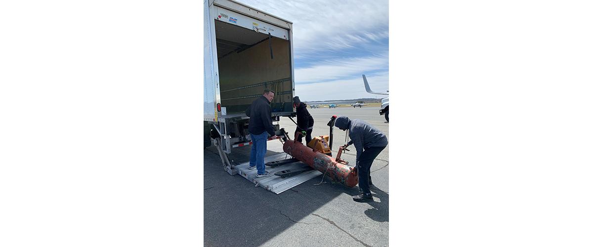 Three people load an orange metal tube onto a truck. 