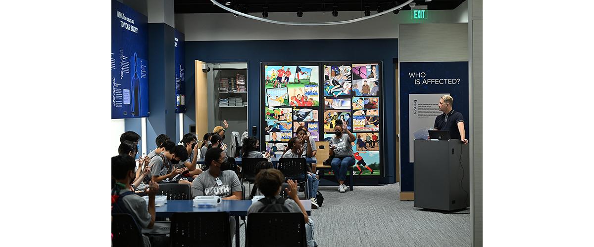 A group of students seated at tables listen to a presenter at the front of the room. 