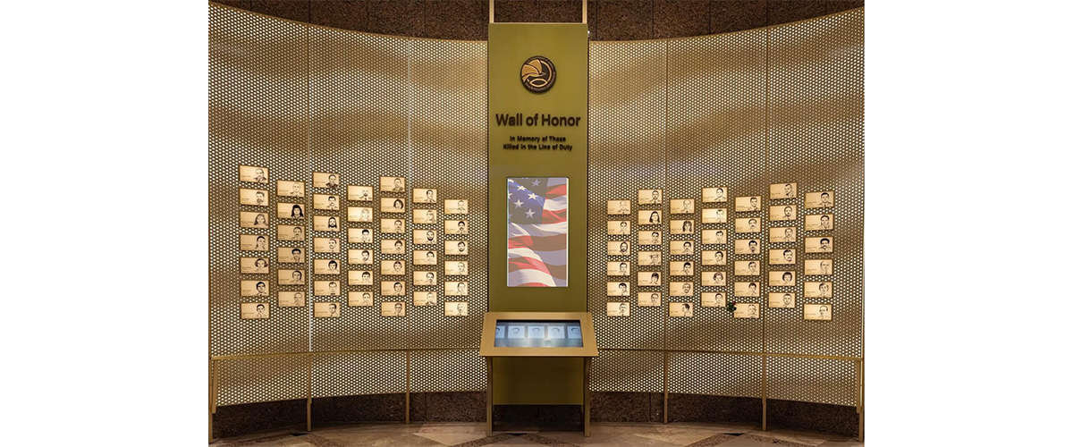 A bronze memorial wall with plaques, each detailing a person's name, title, death date, and picture.