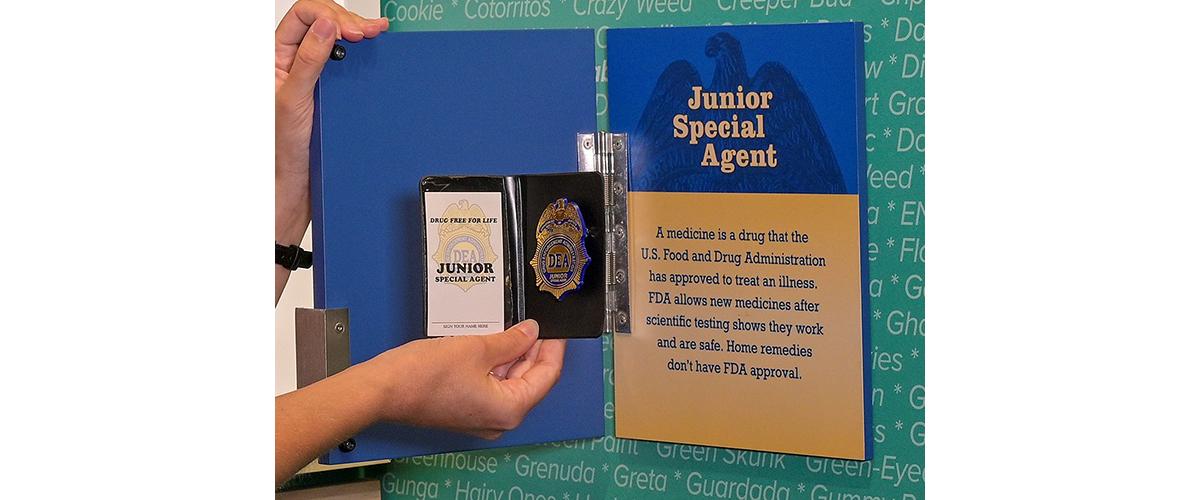 A person holds a badge in front of a museum panel labeled "Junior Special Agent."